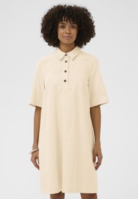 Béžové šaty s krátkym rukávom a golierom, s gombíkovou légou a voľným strihom. Hladká tkanina s jemnou textúrou.