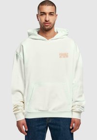 Creme hoodie med en frontlomme og blødt, struktureret stof. Ordet "SPRING" er trykt i ferskenfarve på venstre brystområde.