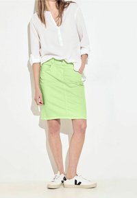 Falda de mezclilla verde claro con corte recto y bolsillos frontales, combinada con una camisa blanca de botones y zapatos blancos con detalles en negro.