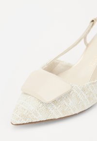 Zapato slingback de punta afilada en material texturizado color crema con un acento liso color beige en la parte superior. Presenta una correa delgada y un suave forro interior.