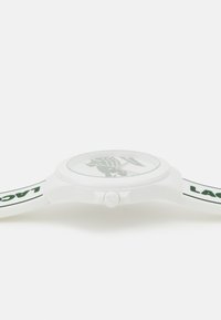 White silicone watch with a round face, green graphic design, and "LACOSTE" printed in green on the strap. Features a side crown.