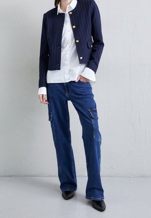Osoba ubrana w granatową marynarkę w prążki, skróconą, z złotymi guzikami, białą koszulę z kołnierzykiem, niebieskie dżinsy typu cargo i czarne spiczaste buty.