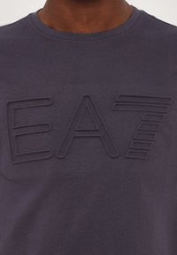 Tamsiai mėlyna marškinėliai su trikotažine apykakle. Priekyje yra tekstūruotas, toninis embosuotas "EA7" logotipas. Minkšta medvilnės medžiaga.