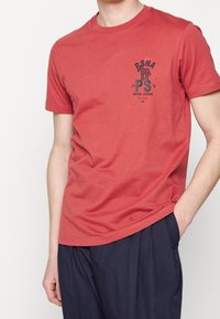 Camiseta de algodón roja con cuello redondo, mangas cortas y un diseño gráfico impreso que presenta texto y un motivo de esqueleto en el pecho.