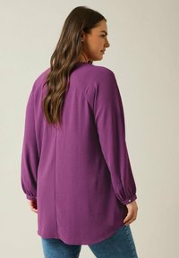 Blusa viola con maniche lunghe, tessuto strutturato e orlo curvo. Presenta dettagli con bottoni sulle maniche e una vestibilità rilassata.