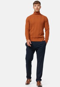Pull à col roulé en orange brûlé texturé avec un motif en zigzag, associé à un pantalon bleu marine et des chaussures en cuir marron.