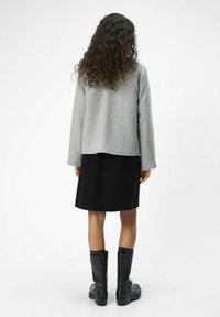 Szary, wełniany płaszcz, czarna spódnica do kolan, czarne wysokie buty, widoczne teksturowane loki, widok z tyłu. Minimalistyczny design, czyste linie.