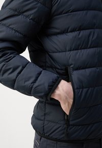 Navy blue puffer jacket with horizontal quilted pattern, zippered pocket on the side, and elastic cuffs; lightweight and insulated design.