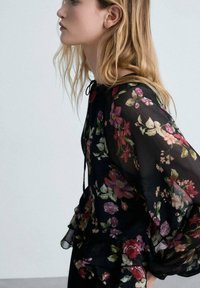 Blusa nera trasparente con stampa floreale nei toni di rosso, rosa e verde. Maniche con bordi arricciati e dettaglio con cordoncino al collo.
