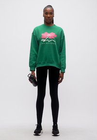 Γυναίκα όρθια, φοράει πράσινο φούτερ Quiksilver, μαύρο κολάν, μαύρα αθλητικά παπούτσια, κρατώντας μαύρο μπουκάλι νερού στο δεξί χέρι.