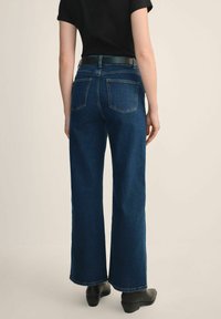 Osoba ubrana w ciemnoniebieskie, szerokie dżinsy z wysokim stanem, z tylnymi kieszeniami, czarny pasek, czarną koszulę z krótkim rękawem i czarne botki za kostkę.