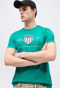 Νέος άνδρας που φοράει πράσινο t-shirt GANT με την επιγραφή "American Sportswear" και μαύρο καπέλο μπέιζμπολ, με σταυρωμένα χέρια, κοιτώντας προς τα πλάγια.