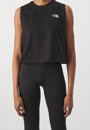 Person iført en sort ærmeløs crop top med The North Face-logo og højtaljede sorte leggings mod en ensfarvet baggrund.