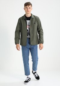 Veste légère vert olive avec des boutons-pression, manches flottantes, superposée sur un t-shirt graphique noir. Associée à un jean bleu et des baskets noires.