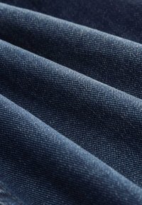 Denimová tkanina, tmavě modrá, s viditelnou texturovanou vazbou a jemným vyblednutím, složená v vrstvených, šikmých sekcích, které ukazují hloubku a detail.