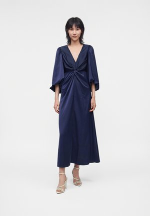 Vrouw die een lange marineblauwe jurk draagt met pofmouwen, een diepe V-hals en een gedraaid knoopdetail, gecombineerd met witte sandalen met hoge hakken.