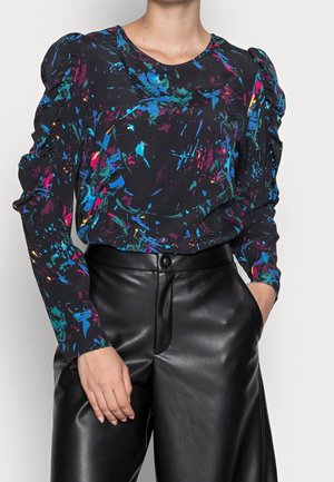Femme portant un chemisier noir à manches longues avec des motifs abstraits colorés et des épaules bouffantes, associé à un pantalon en cuir noir taille haute.