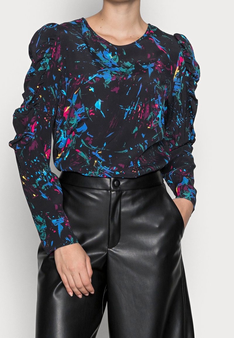 Femme portant un chemisier noir à manches longues avec des motifs abstraits colorés et des épaules bouffantes, associé à un pantalon en cuir noir taille haute.