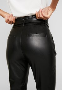 Czarne spodnie z eko skóry z wysokim pasem, wyposażone w dwie tylne kieszenie i gładką, matową fakturę. Zbliżenie na linię talii.