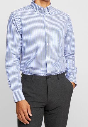 Homme portant une chemise boutonnée à rayures verticales bleues et blanches et un pantalon gris foncé, une main dans la poche et l'autre détendue le long du corps.