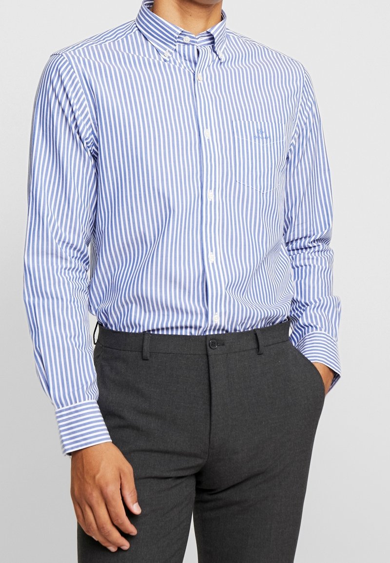 Homme portant une chemise boutonnée à rayures verticales bleues et blanches et un pantalon gris foncé, une main dans la poche et l'autre détendue le long du corps.