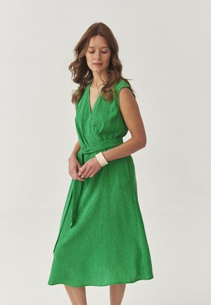 Green wrap dress with a V-neckline, short sleeves, and a textured fabric. Features a waist tie and a flowing mid-length skirt.
