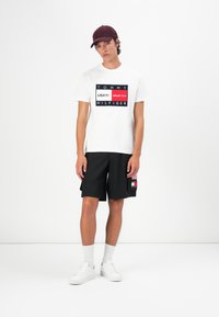 T-shirt blanche avec imprimé graphique bleu marine et rouge, shorts noirs avec patch logo, baskets blanches et casquette bordeaux, se tenant sur un fond uni.
