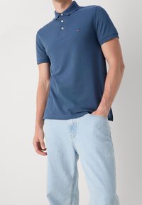 Homme portant un polo bleu à manches courtes et un jean bleu clair avec une main dans la poche sur un fond uni.