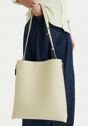 Person trägt dunkelblaue Hose und hellgelbe Bluse und hält große beige Schultertasche mit verstellbarem Riemen.