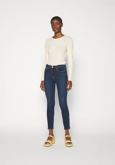 Haut à manches longues crème clair, jean skinny bleu foncé et sandales à talons jaunes avec des brides à la cheville. Design simple et ajusté, textures lisses.