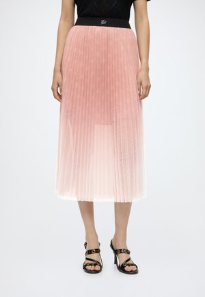Frau trägt ein schwarzes Oberteil, einen rosa plissierten Midi-Rock mit Farbverlauf und schwarze High-Heel-Sandalen mit goldenen Akzenten, steht aufrecht.