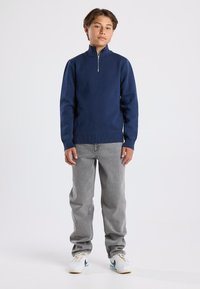 Laivastonsininen vetoketjullinen puolipaita, teksturoitu neulemateriaali, korkea kaulus, yhdistetty vaaleanharmaisiin farkkuihin ja valkoisiin lenkkareihin mustilla yksityiskohdilla.