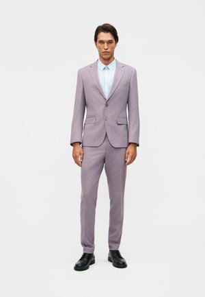 Άνδρας όρθιος που φοράει ανοιχτό μοβ κοστούμι, γαλάζιο πουκάμισο με γιακά και μαύρα επίσημα παπούτσια, μπροστά σε λευκό μονόχρωμο φόντο.