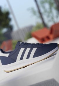 Zapatilla Adidas azul marino con rayas blancas, suela beige y diseño con cordones, exhibida al aire libre sobre una superficie blanca.