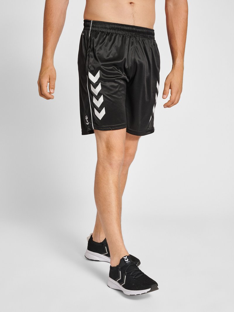 Mann trägt schwarze Sportshorts mit weißen Chevron-Streifen und schwarze Sportschuhe und steht vor einem einfarbigen Hintergrund.