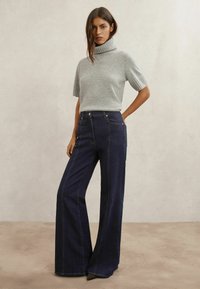 Maglione grigio a maniche corte con collo alto abbinato a pantaloni scampanati in denim scuro a vita alta, con tasche frontali e dettagli di cuciture visibili.