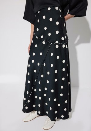 Zwarte maxi rok met grote witte polka dots, asymmetrische naaddetails, gedragen met een zwart topje en witte veterschoenen.