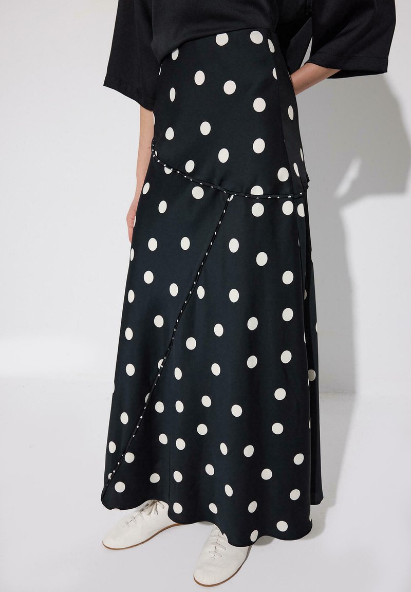 Black maxi skirt with large white polka dots, asymmetrical seam details, worn with black top and white lace-up shoes.