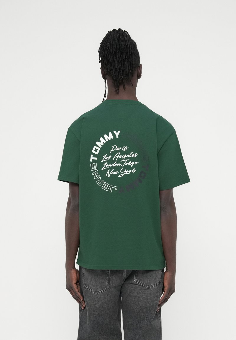 Πράσινο βαμβακερό t-shirt με το λεκτικό «TOMMY JEANS» τυπωμένο σε λευκό και μαύρο στην πλάτη, με ονόματα πόλεων σε κυκλική διάταξη. Κοντά μανίκια.