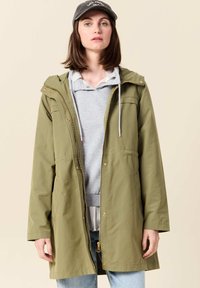 Olivgrön parkajacka med huva, framfickor, guldfärgade dragkedjor och justerbara snoddar, lagd över en grå sweatshirt.