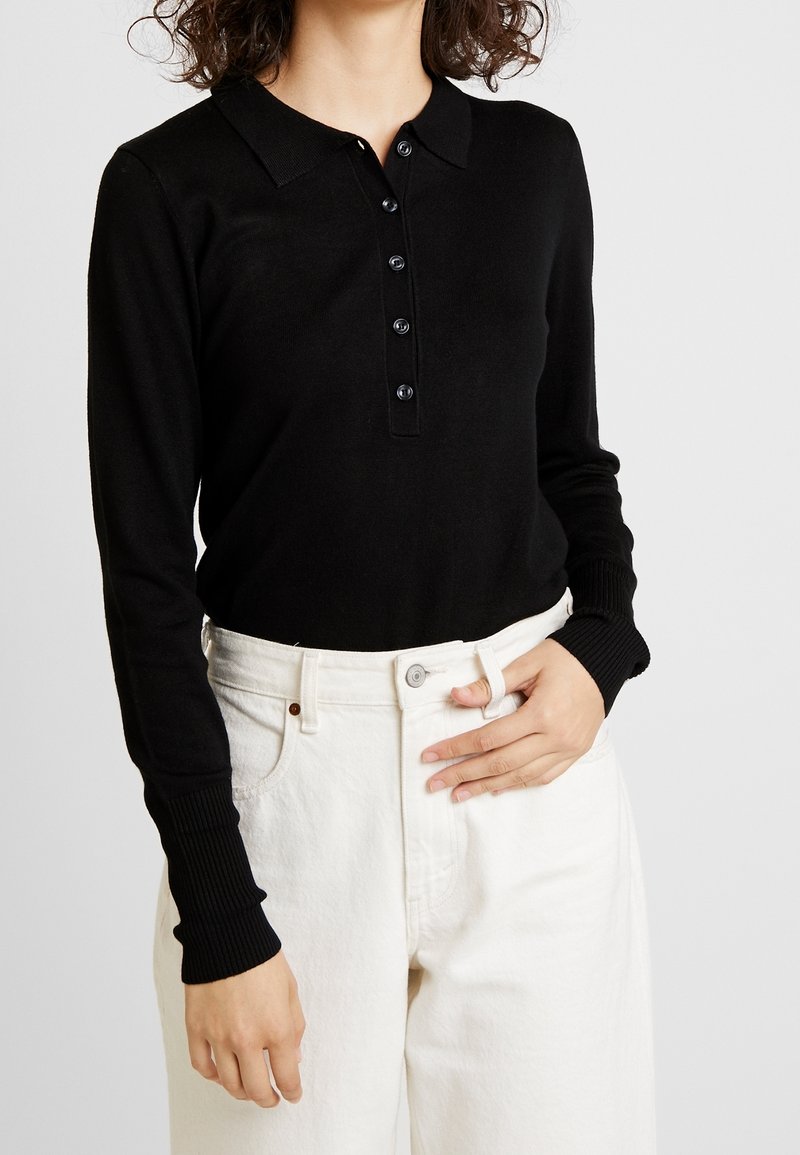 Zwart poloshirt met lange mouwen en een kraag, voorzien van een knoopsluiting aan de voorkant en geribbelde manchetten, gecombineerd met witte jeans met hoge taille.