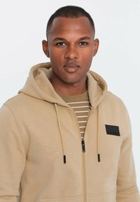 Sudadera beige con tela texturizada, cierre frontal y parche de logo negro. Debajo, se ve una camiseta a rayas en tonos beige y marrón claro.
