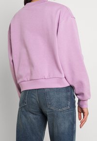 Felpa lilla con scollo rotondo, polsini e orlo a costine; presenta una vestibilità comoda e un tessuto morbido. Abbinata a jeans blu.