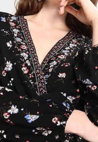 Blusa nera floreale con scollo a V profondo e maniche a puffo. Il design include piccoli motivi floreali colorati e cuciture decorative.