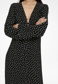 Schwarzes Polka-Dot-Kleid mit tiefem V-Ausschnitt und langen Ärmeln, das einen vorderen Verschluss und einen seitlichen Schlitz für zusätzlichen Detailreichtum aufweist.