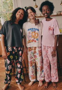 Troje dzieci nosi koszulki z nadrukami i wzorzyste piżamowe spodnie. Materiały są bawełniane, zdobione kolorowymi motywami jedzenia w różnych kolorach.