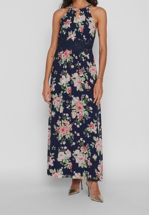 Vrouw draagt een marineblauwe mouwloze maxi-jurk met roze en witte bloemen, kantdetail in de taille en gouden hoge hakken.