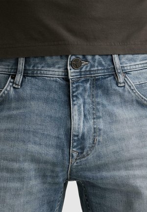 Close-up af taljen på lyseblå, falmede denimjeans med knap og lynlås, båret sammen med en indstoppet mørkebrun skjorte.