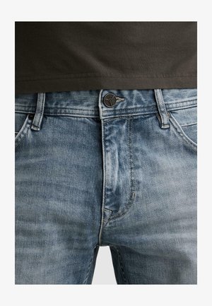 Close-up af taljen på lyseblå, falmede denimjeans med knap og lynlås, båret sammen med en indstoppet mørkebrun skjorte.