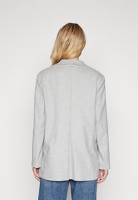 En blond person klädd i en ljusgrå blazer och blå jeans, står med ryggen mot kameran mot en enkel vit bakgrund.